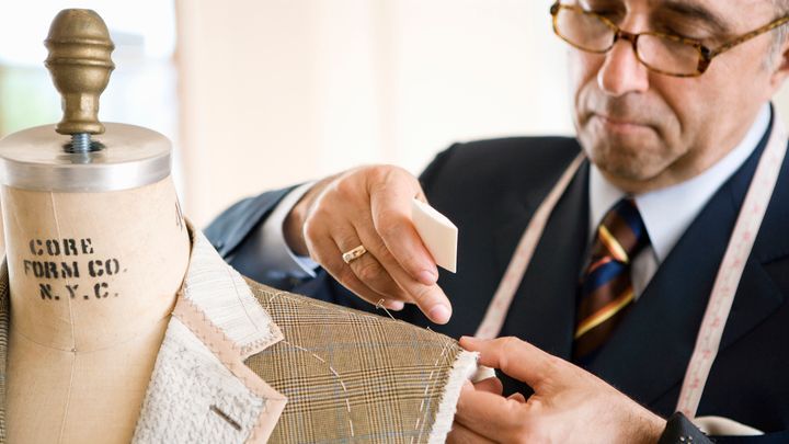 Medium closeup of 55yr Cauc male bespoke tailor in dark suit top of head out of frame making chalk marks on shoulder of...