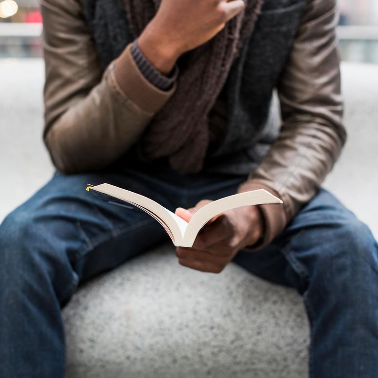 Vista parcial de un joven sentado en un banco leyendo un libro; concepto de los mejores libros que todo hombre deber leer.