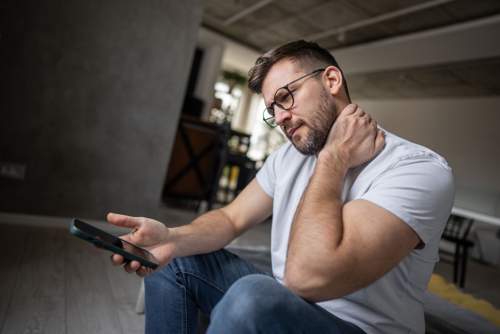 Mid adult man sitting on the floor holding a smartphone and experiencing neck pain from poor posture reflecting the...